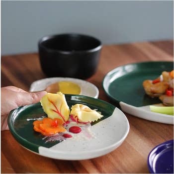 A hand holds a green and white plate with ravioli, garnished with vegetables and sauce. In the background, there are other plates with food and a black cup