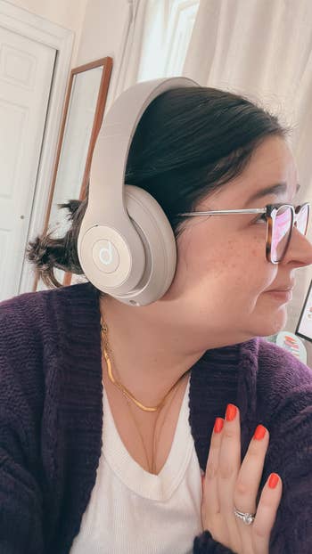 Person wearing headphones and glasses, looking away; wears a white shirt and purple cardigan, showcasing jewelry