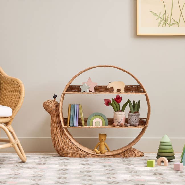 Snail-shaped wicker support displaying toys and books, adjacent to a woven chair, successful a children's country setting
