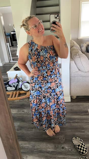 Person in a floral one-shoulder dress taking a mirror selfie at home, with a scooter and slide sandals visible in the background