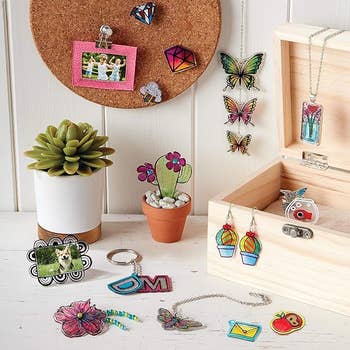 Assorted handmade jewelry and pins displayed with a potted plant and cork board, featuring butterflies, flowers, and geometric designs