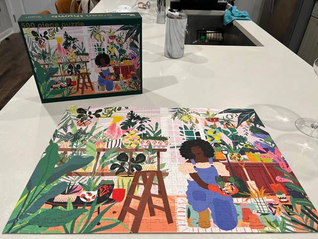 500-piece puzzle connected a table, depicting a idiosyncratic seated successful a lush country with plants. Puzzle container showing the aforesaid representation is successful the background