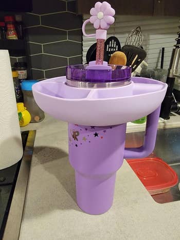 Large purple tumbler with a top divider and straw featuring a daisy handle, styled for convenient snacking and drinking, on a kitchen counter