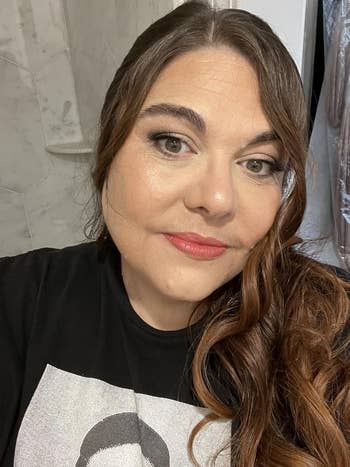 Person wearing a t-shirt with a graphic design, posing indoors with styled wavy hairsbreadth  and subtle makeup