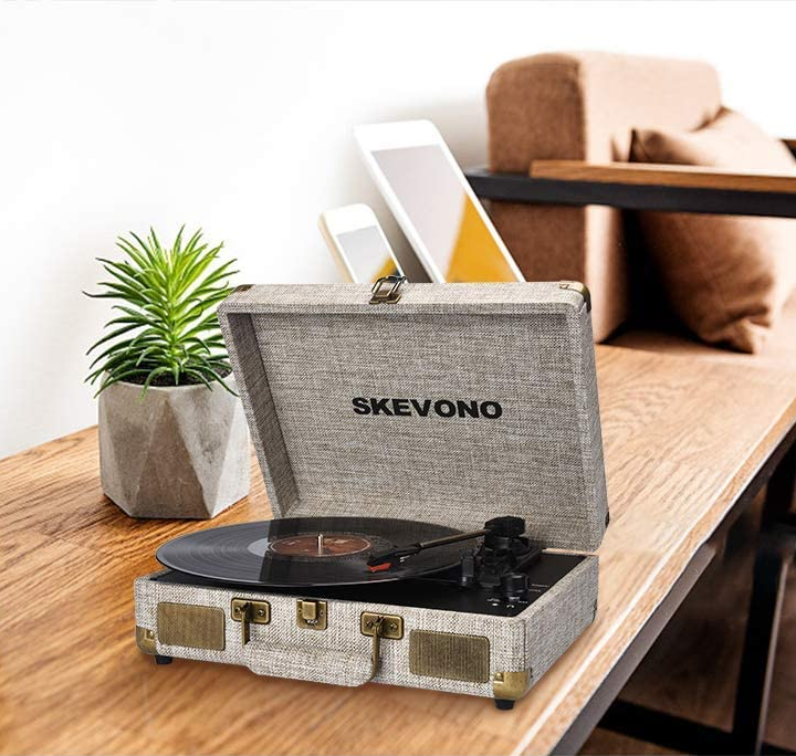 Gray portable suitcase turntable with black stylus on vinyl sitting on top of wooden table next to plants