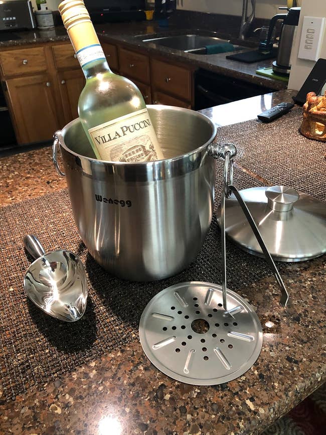 Wine bottle in a stainless steel ice bucket with a lid, strainer, and scoop on a kitchen counter