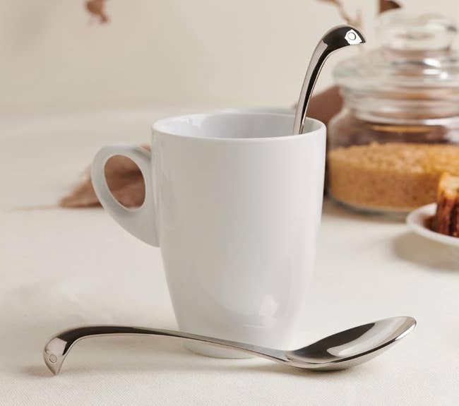 A silver spoon with a design on the top of it to look subtly like the loch ness monster is popping out of a mug 