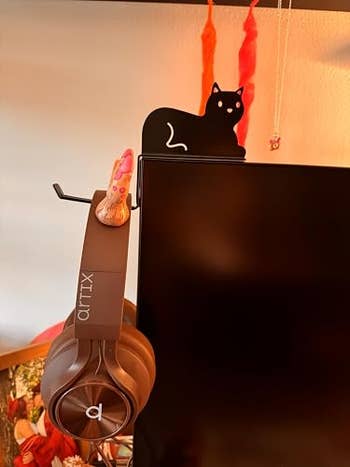 Headphones hanging on a monitor featuring a black cat-shaped topper and a small decorative shoe