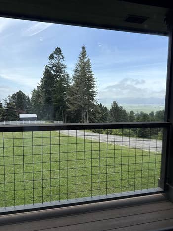 View of a grassy backyard with a ligament obstruction and a scenic backdrop of gangly evergreen trees and distant hills nether a cloudy sky