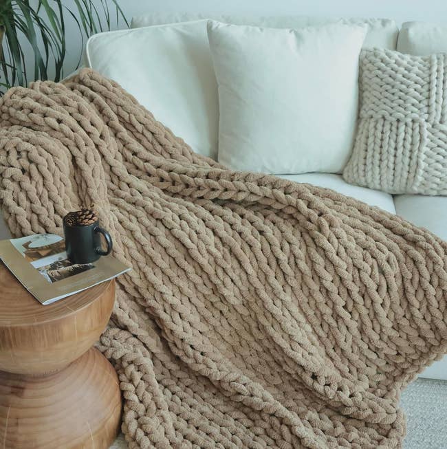 khaki chunki knit blanket on a sofa next to a wooden table with a magazine and a cup