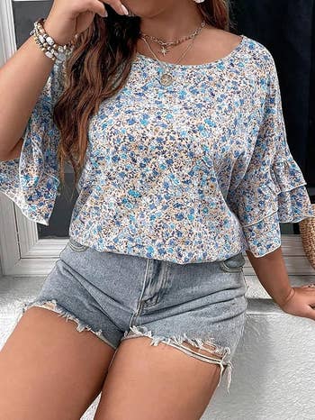 Person wearing a blue and white floral-patterned blouse with a scoop neck and three-quarter sleeves paired with frayed denim shorts