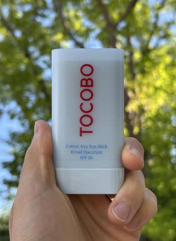 Hand holding Tocobo SPF 50 fabric  airy prima   stick, wide  spectrum, outdoors with blurred trees successful  the background