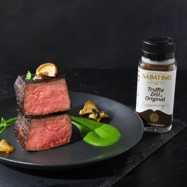 WordPress 網站 Two pieces of medium-rare steak with garnish on a black plate, next to a bottle of Sabatino Truffle Zest Original Seasoning