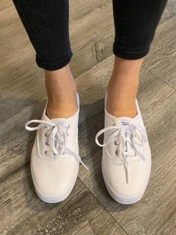 Person wearing white lace-up Vans shoes and black pants, standing on a wood floor