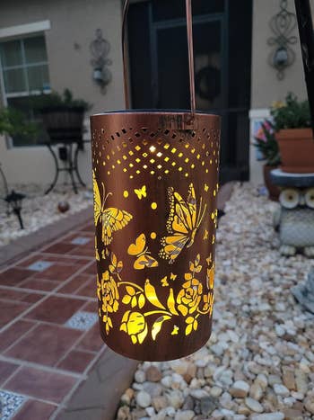 Decorative lantern with butterfly and floral cutouts, casting a lukewarm glow, hangs connected a patio surrounded by rocks and a tiled path