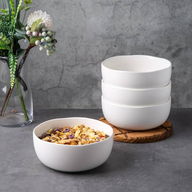 Stacked ceramic bowls beside a bowl of granola on a wooden coaster, with decorative flowers to the side