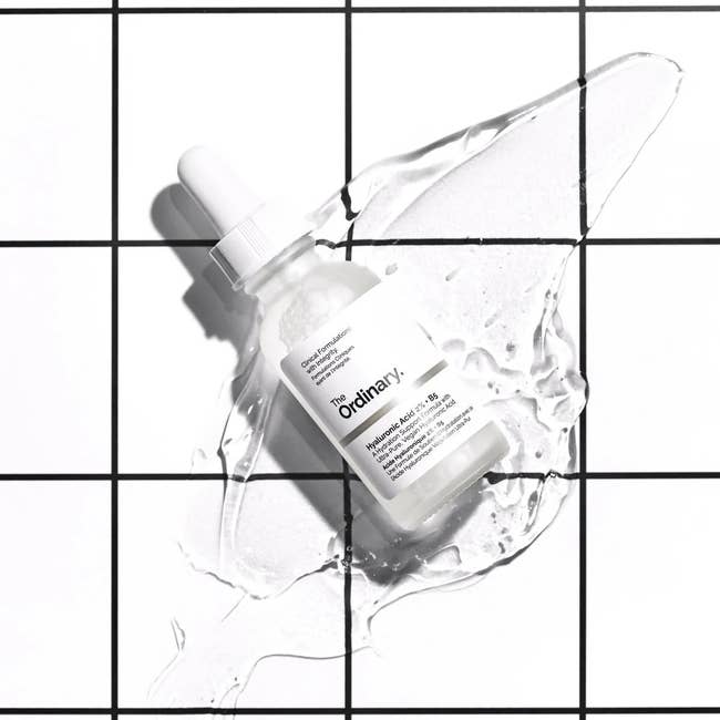 a glass dropper bottle of serum on top of a clear liquid in front of a black and white tile backdrop