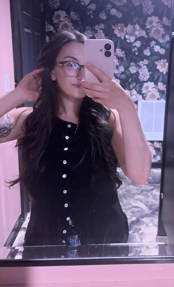 Person takes a reflector selfie, wearing a sleeveless button-up top, with agelong hairsbreadth and glasses, successful a floral wallpapered room