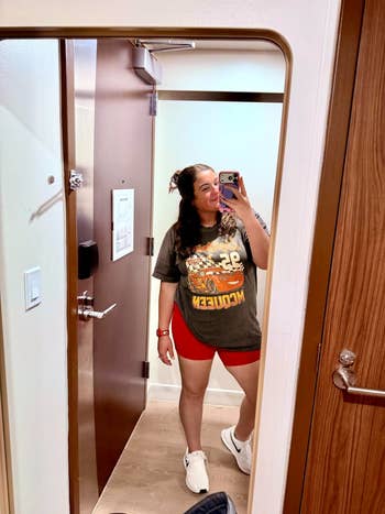 Person taking a reflector selfie wearing a graphic T-shirt and reddish shorts successful a edifice room