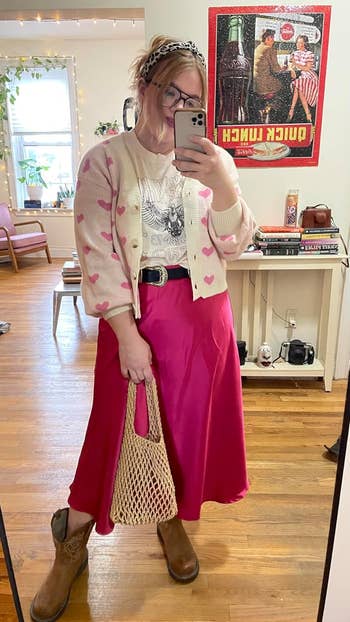 Person in a patterned cardigan, graphic tee, pink skirt, with a netted bag and cowboy boots, taking a mirror selfie