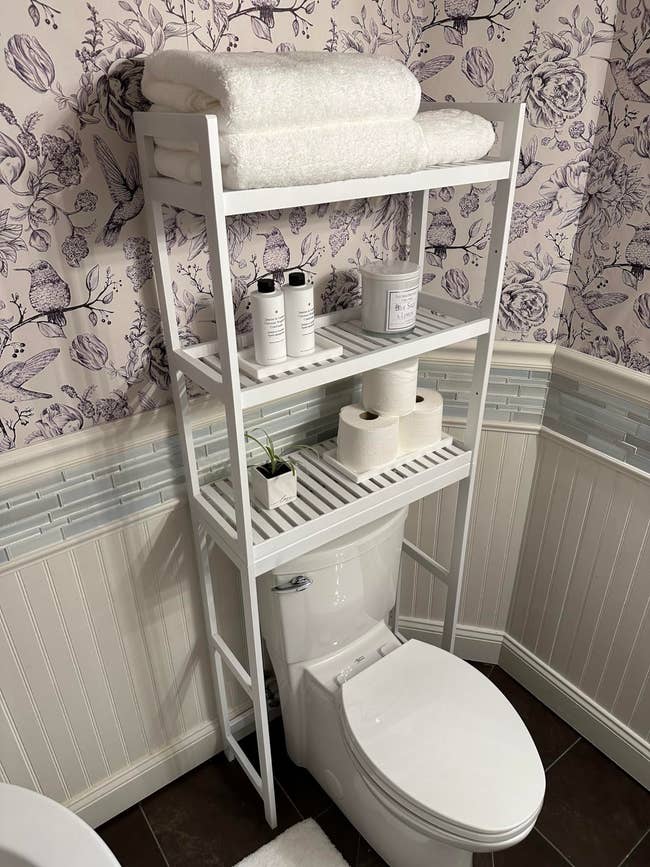 Bathroom shelving unit over toilet with towels, toiletries, and toilet paper on shelves