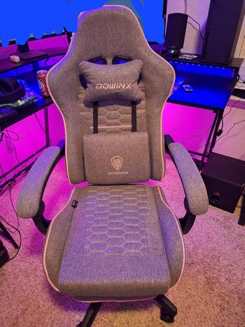 Gaming chair with ergonomic design and lumbar support, suitable for office or gaming setups. Placed in a room with tech equipment and shelves