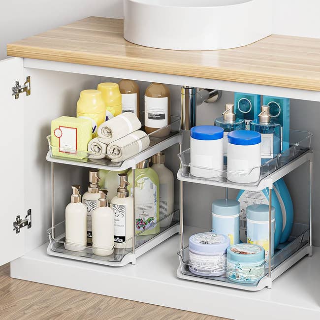 Organized under-sink cabinet with assorted toiletries, towels, and cleaning supplies neatly stored on tiered trays. Perfect for home organization