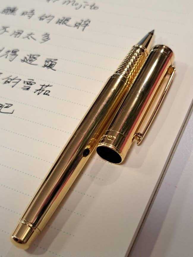 A gold pen with its cap placed beside it rests on an open notebook with handwritten text in a language other than English