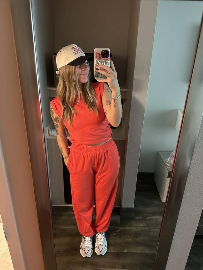 Person taking a reflector  selfie wearing a casual, sleeveless jumpsuit, sneakers, and a cap, holding a smartphone successful  1  hand