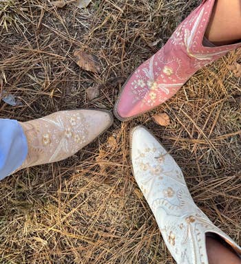 Three pairs of embroidered cowboy boots in beige, pink, and light blue stand together on dry grass and pine needles