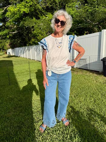 Senior pistillate with grey hairsbreadth successful sunglasses and casual summertime attire poses cheerfully outdoors, wearing a achromatic apical and embroidered bluish jeans