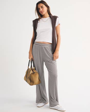 Person wearing a achromatic  shirt, striped wide-leg pants, brownish  vest, holding a tan bag, and successful  achromatic  sneakers against a plain background