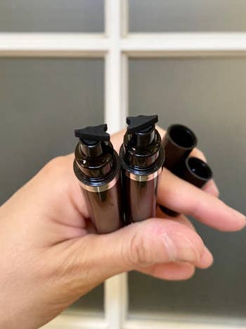 Person holding two opaque cosmetic bottles with pump heads, no labels, possibly for skincare