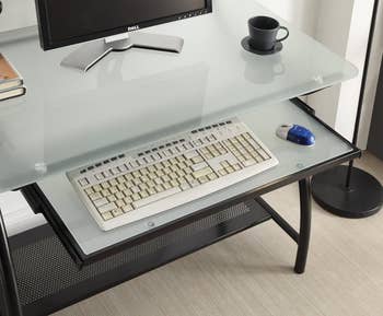 Glass-top desk with a keyboard and blue mouse on a pull-out tray, monitor and books on top, and a coffee cup beside the monitor