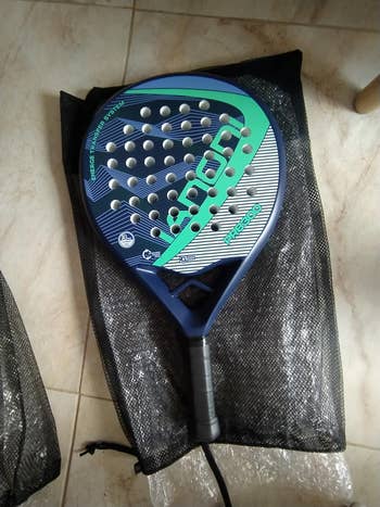 Padel racket with a perforated design, resting on a black mesh bag, photographed on a tiled floor