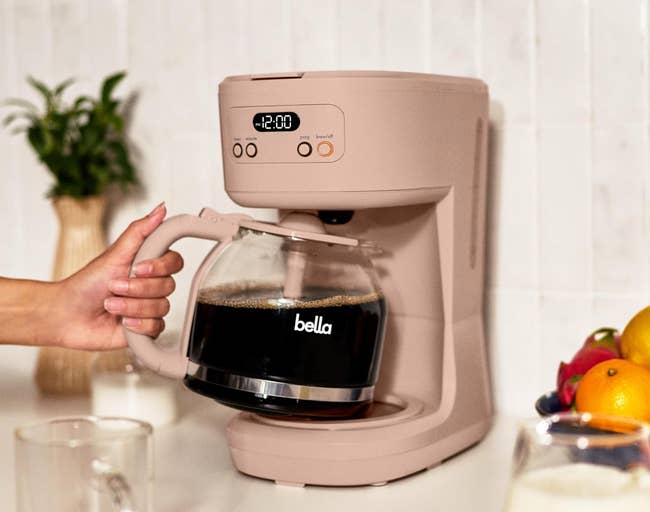 Person pouring coffee from a Bella coffee maker on a kitchen counter, with fruit and a plant in the background