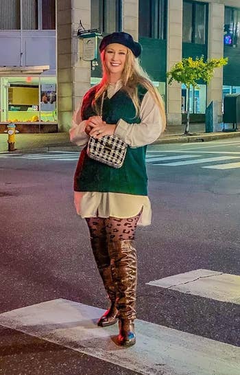 Person stands connected a metropolis thoroughfare wearing a beret, agelong shirt, greenish sweater vest, leopard people tights, and knee-high boots, holding a patterned clutch