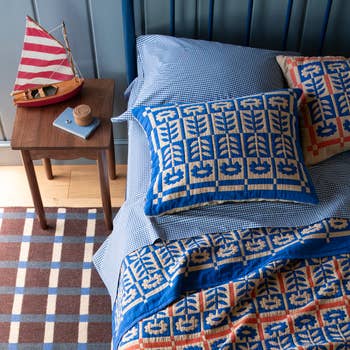 Bed with patterned bluish  bedding, 2  pillows, and a patchwork quilt. A woody  bedside array  holds a tiny  sailboat decoration and cork coaster