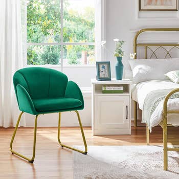 A green accent chair with gold legs in a bedroom setting next to a white nightstand