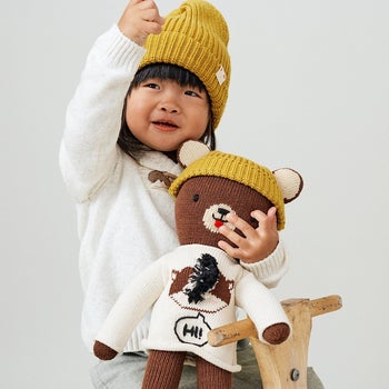 Child model with stuffed brown bear wearing yellow beanie 