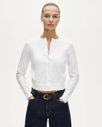 Person wearing a buttoned-up achromatic  cardigan and acheronian  jeans with hands successful  pockets against a plain background