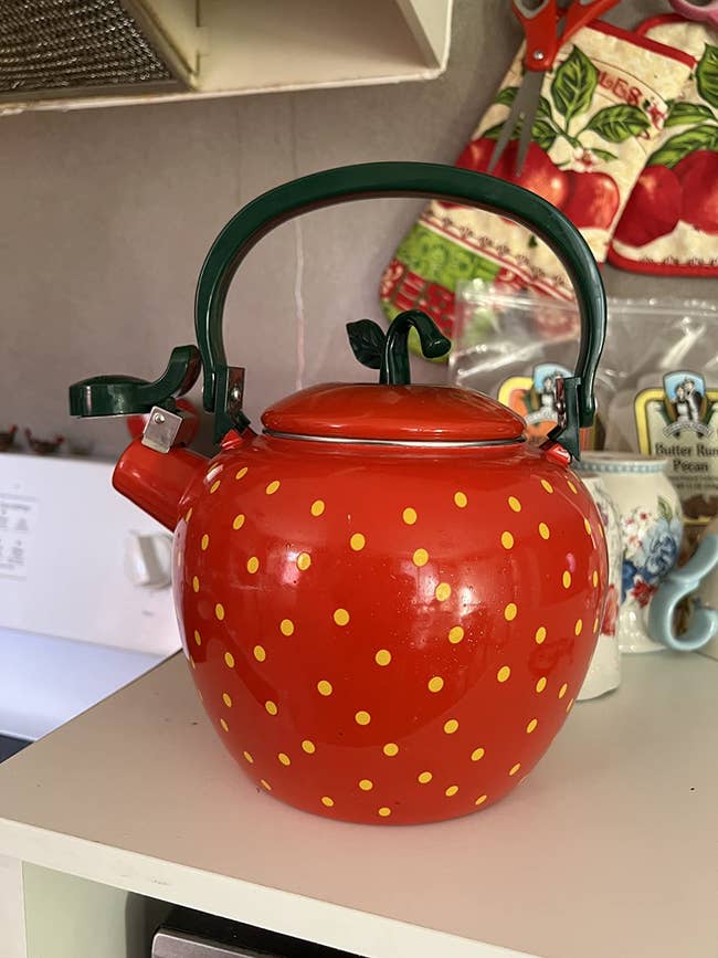 Strawberry-themed teapot with green handle and yellow dots on a kitchen counter. Background includes kitchen items and tea packages
