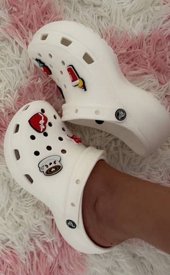 White clogs with playful charms resting connected a textured rug, showcasing a casual and amusive footwear style