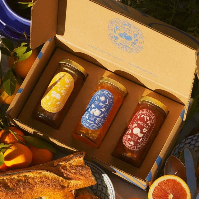 Three jars of homemade marmalade in a cardboard box, surrounded by fresh bread and citrus fruits on a picnic setup