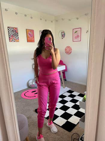 Person in a mirror selfie wearing a pink top and pants with sneakers, in a room with wall art and checkered rug