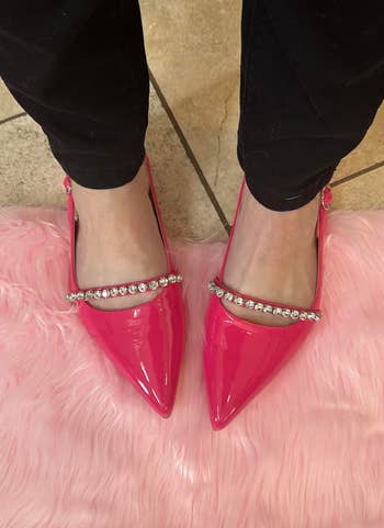 Person wearing shiny pointed shoes with rhinestone straps, lasting connected a pinkish faux fur rug. Black pants are partially visible