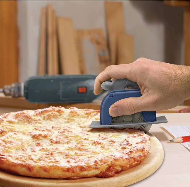 Hand using a circular saw-style pizza cutter on a cheese pizza, with tools and wood in the background