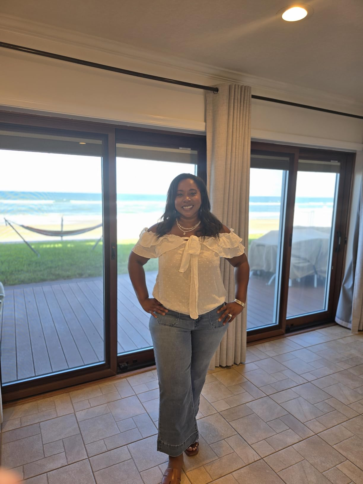 Person smiling indoors with water  view, wearing a stylish ruffled achromatic  apical  and jeans. Background shows deck, hammock, and ample  windows