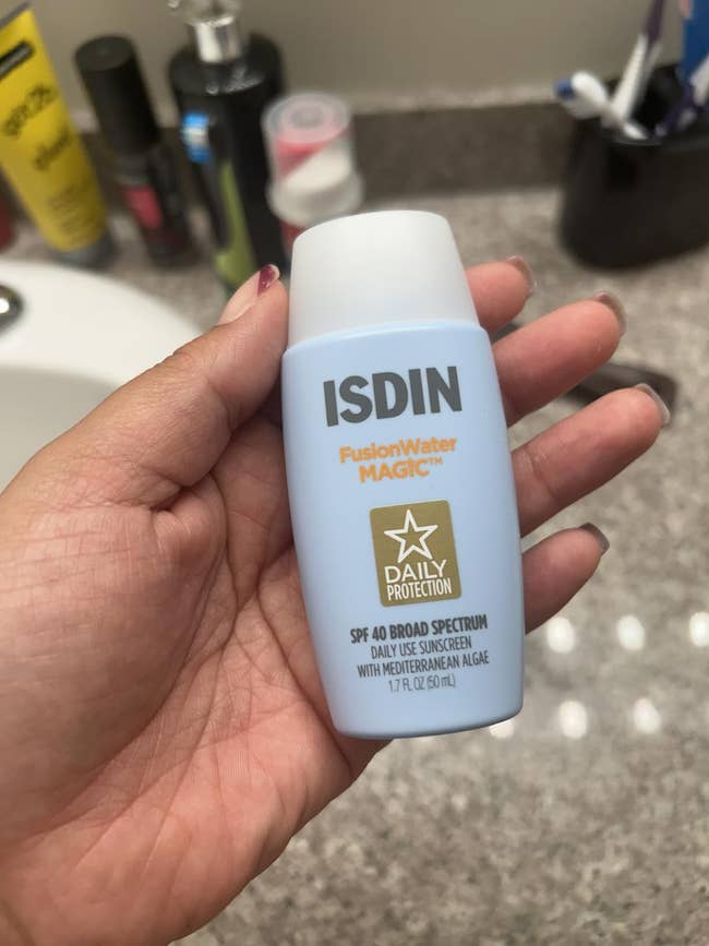 A reviewer's Hand holding ISDIN Fusion Water Magic Daily Protection SPF 40 sunscreen vessel  with disposable   bath  antagonistic  items successful  the background