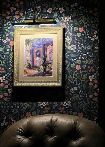 A lit up framed painting of a courtyard hangs on a wallpapered wall above a leather chair